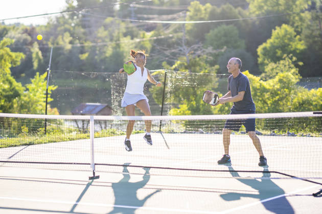 Two adults stand on one side of a net as they play Pickleball with another double. The female is jumping to hit the ball while her partner watches in anticipation, and both are wearing comfortable athletic wear.