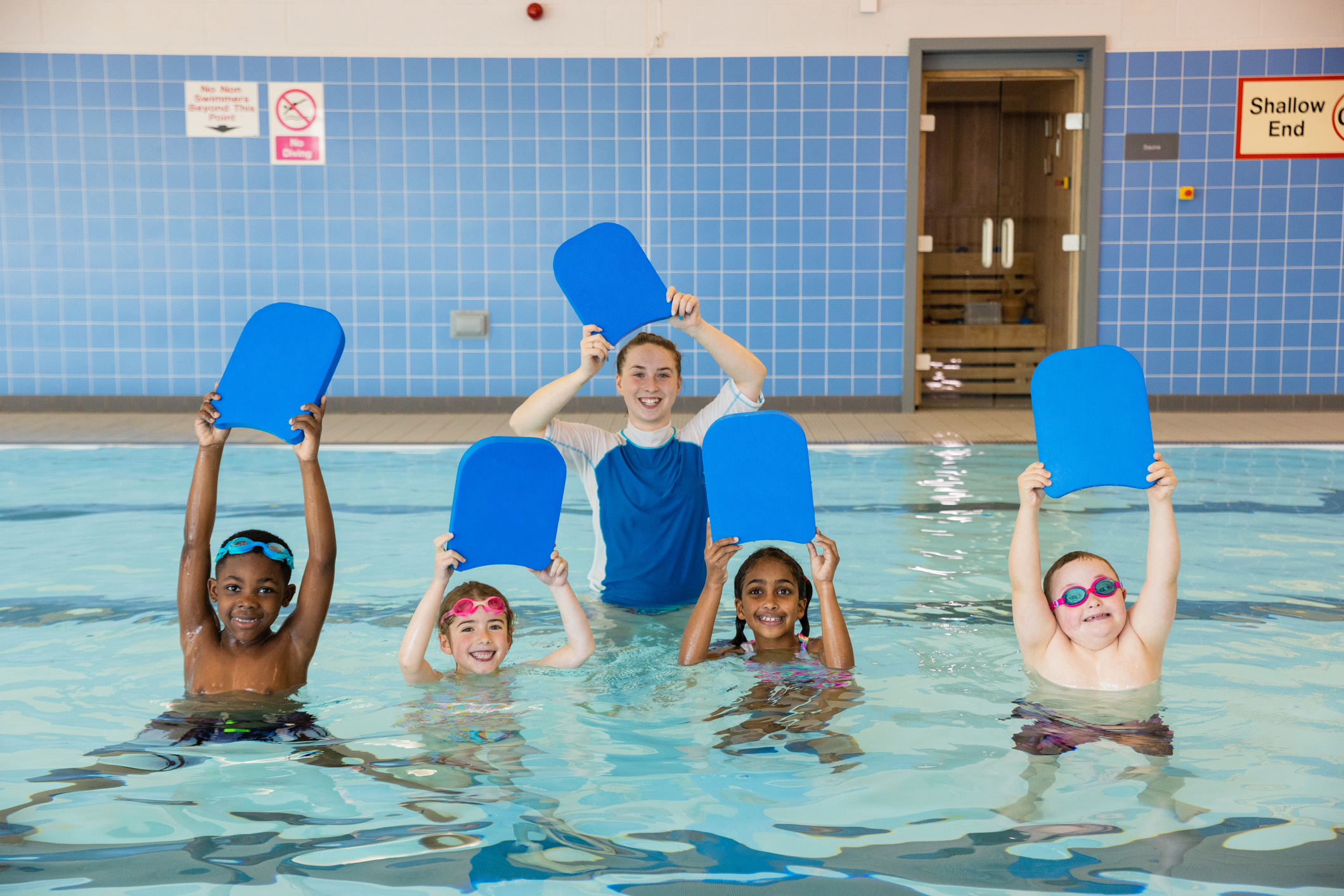 A group of children with their swimming instructor holding swimming floats above their heads in a swimming pool in Boldon, North East England. They are looking at the camera and smiling.
