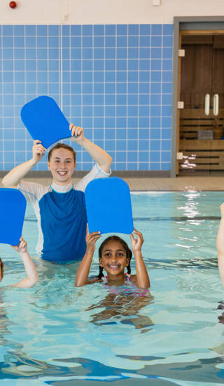 A group of children with their swimming instructor holding swimming floats above their heads in a swimming pool in Boldon, North East England. They are looking at the camera and smiling.