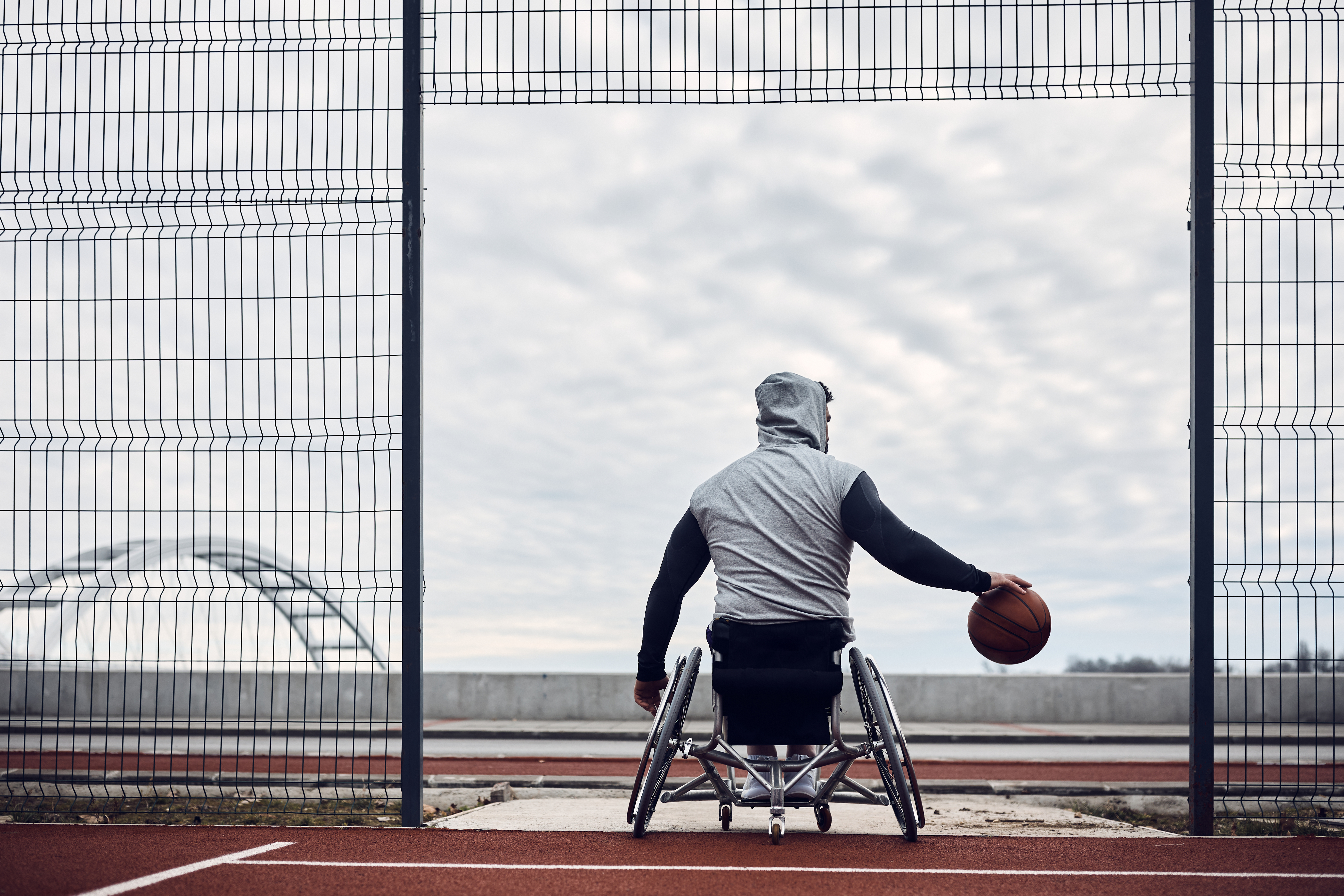 Rug-aanzicht van basketter in rolstoel die dribbelt met bal outdoor, bewolkte lucht 
