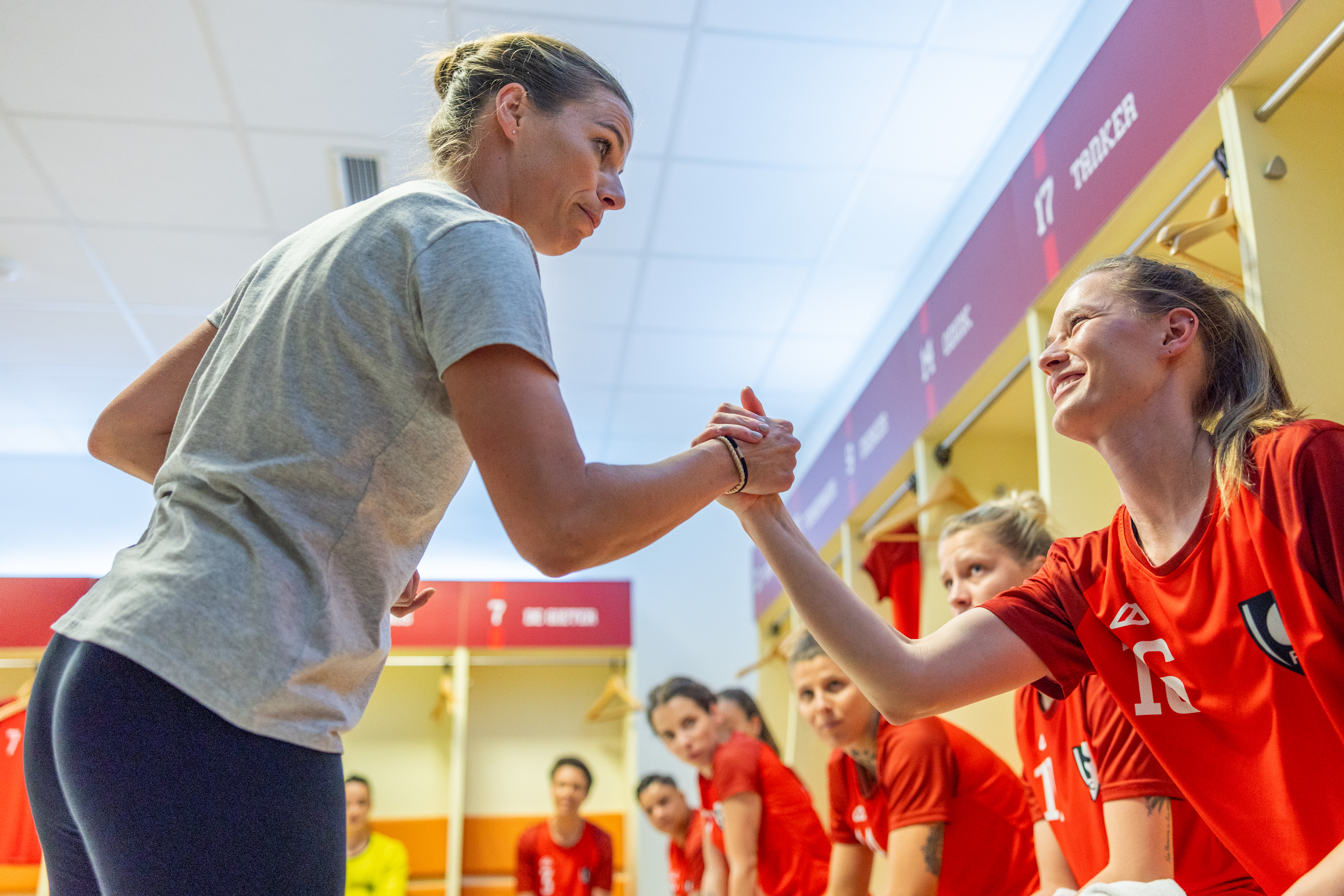 Coach holding hands while talking to smiling female football player sitting in dressing room.