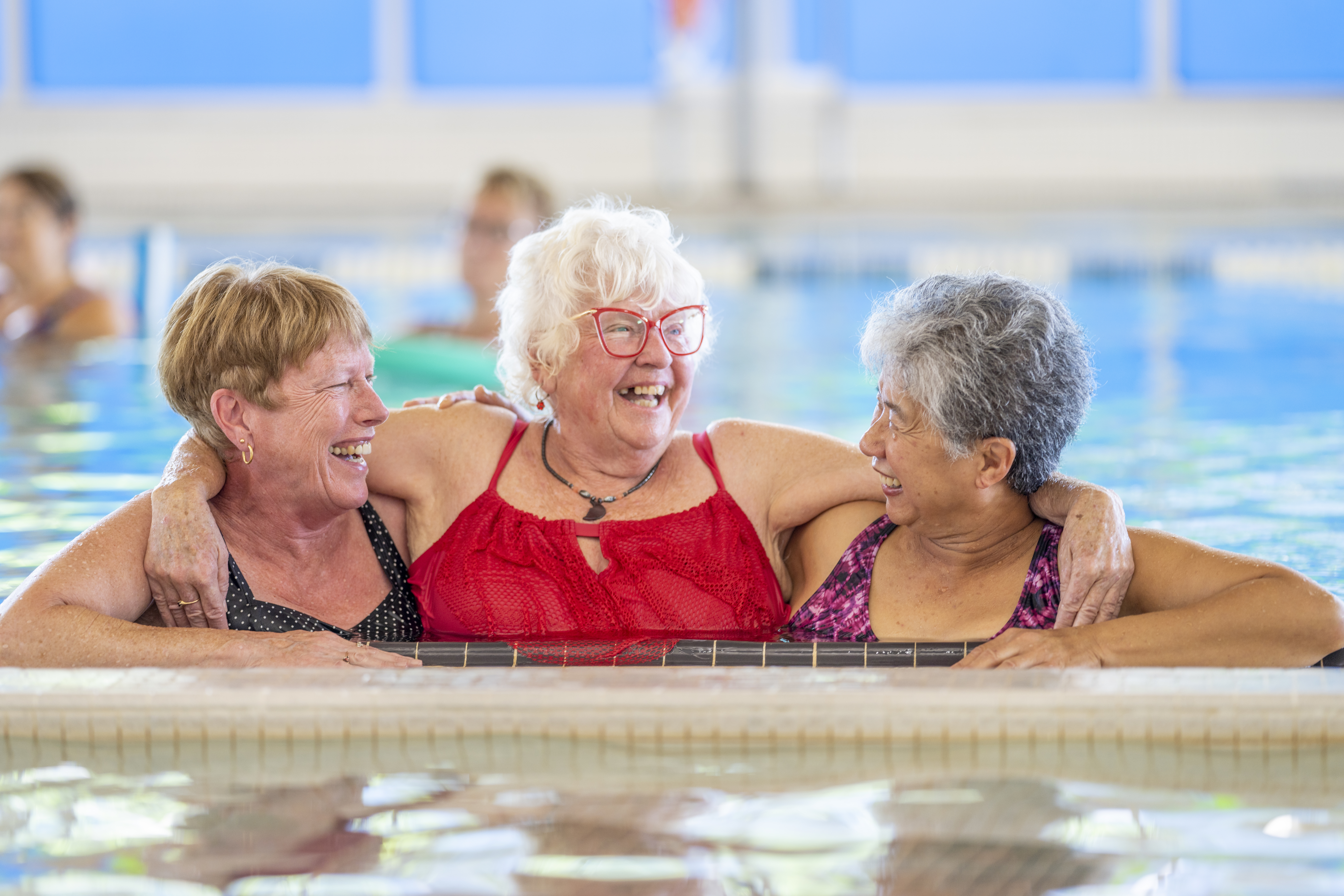 A group of happy seniors taking a water aerobics class at a community centre. The women use various pool equipment to exercise.