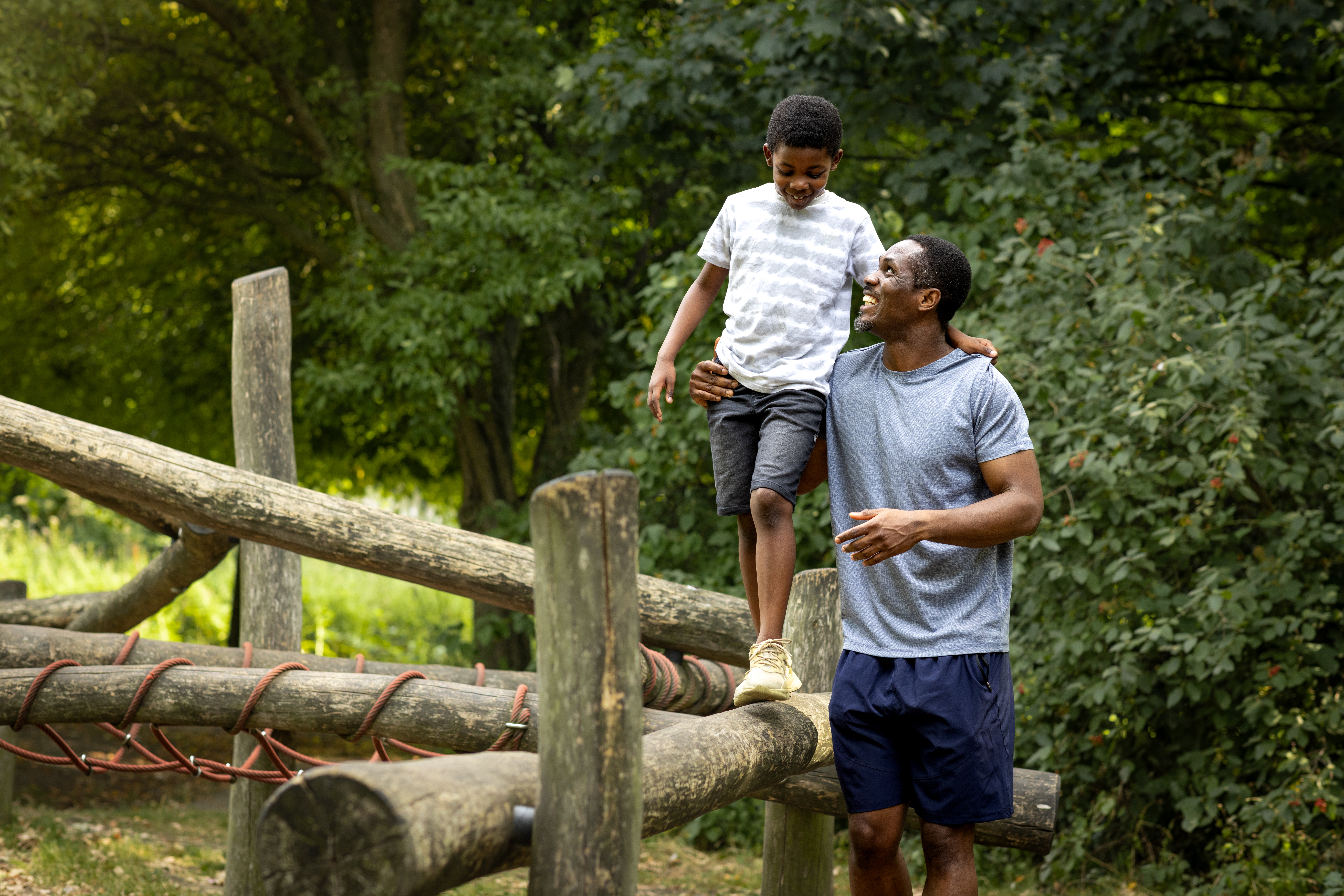 A father and son sending quality time together in their local park. The young boy is balancing on a obsticle course while his dad supports him by the waist. the father is looking up at his son with a smile.