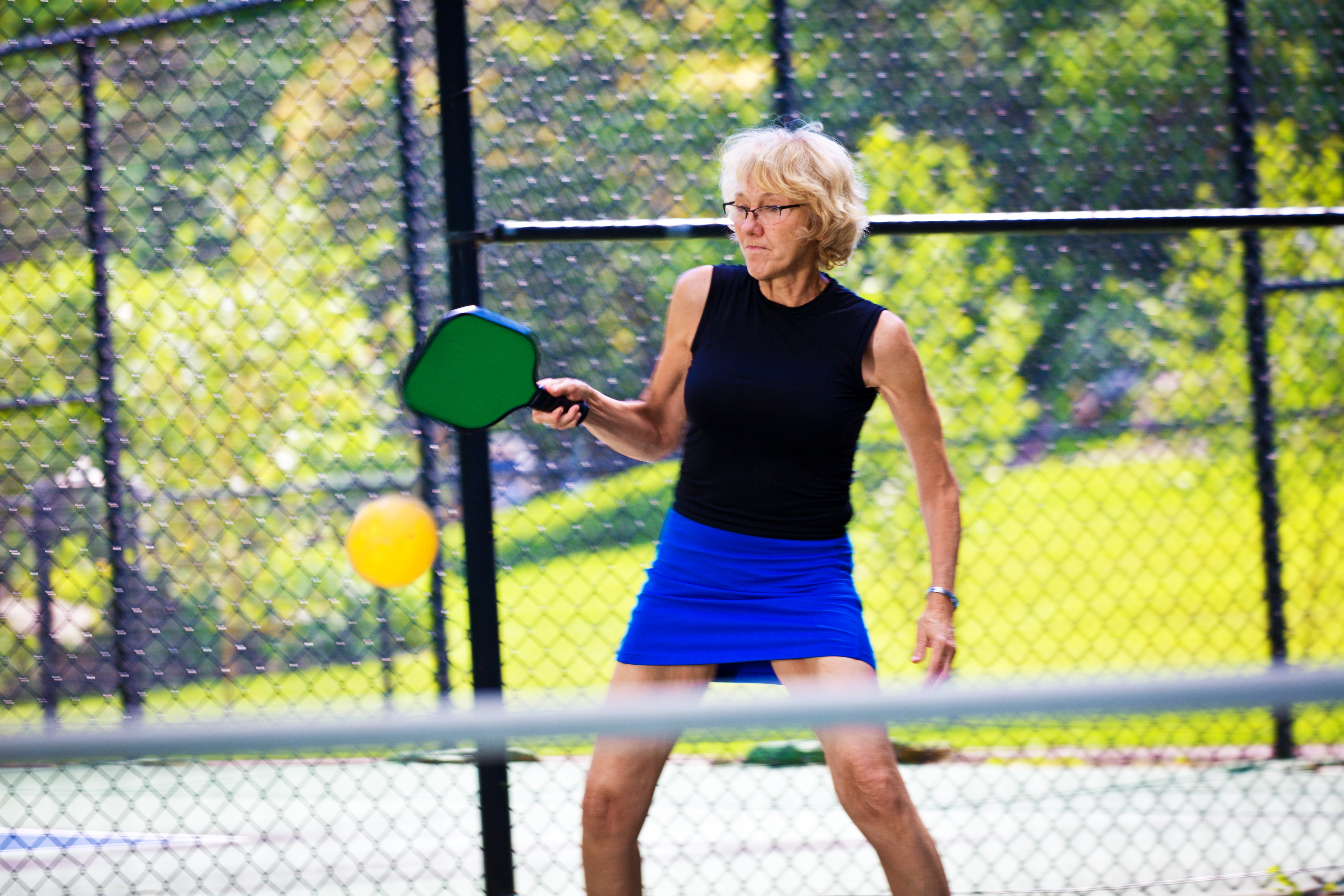 A Caucasian retiree  adult woman pickleball player in action. She is holding a pickleball paddle posing to return the ball in a pickleball court. Photographed in horizontal format with copy space.
