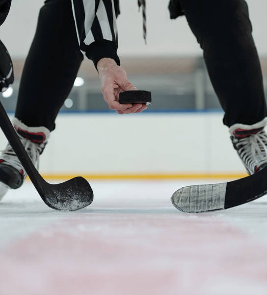 Hand of referee holding puck over ice rink with two players with sticks standing around and waiting for moment to shoot it