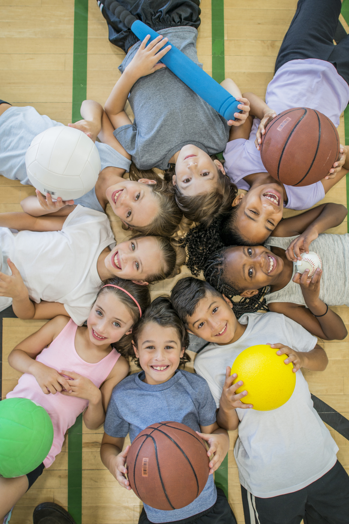 A multi-ethnic group of children are indoors in a health center. They are wearing casual clothing and running shoes. They are lying on the floor with various sports balls, and smiling up at the camera.