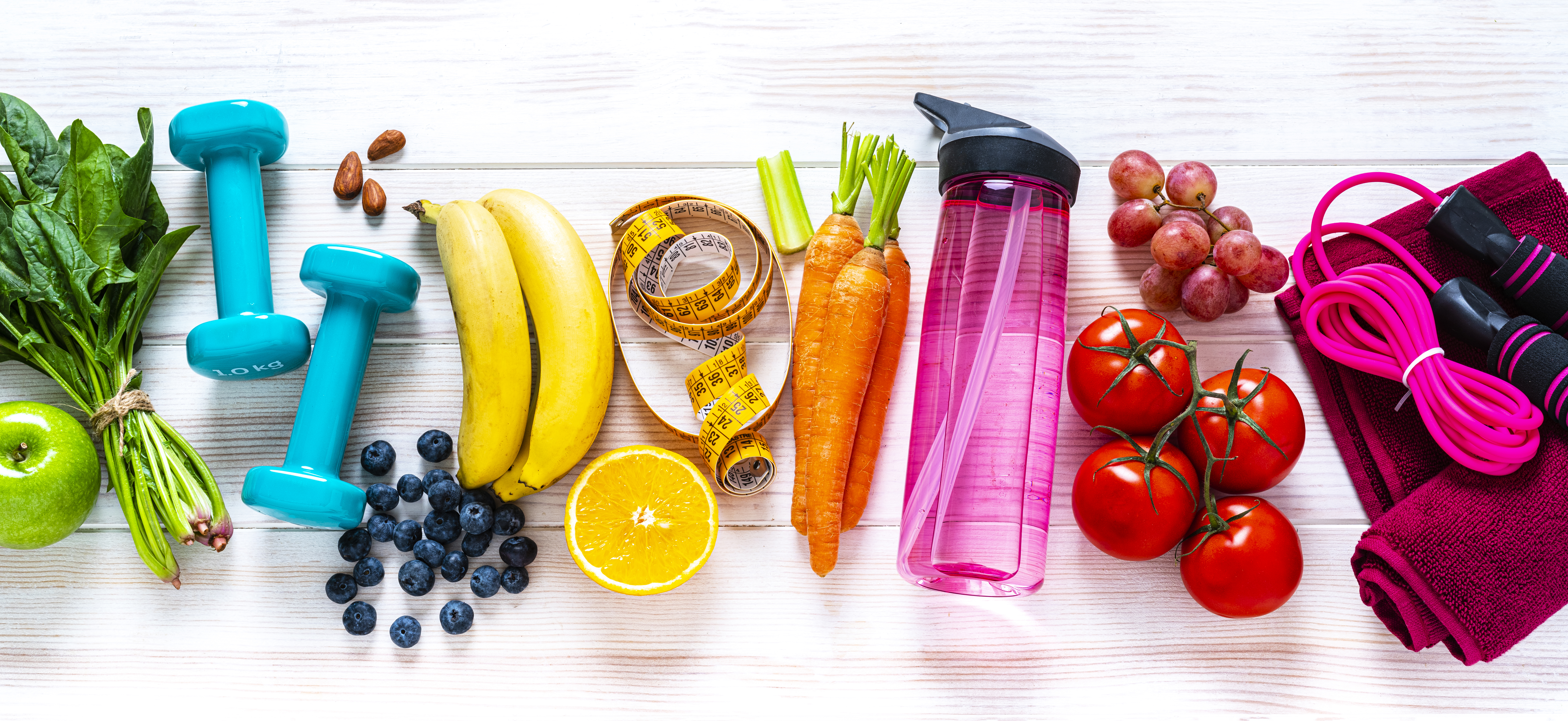 Exercising and healthy eating concept: overhead view of rainbow colored dumbbells, jump rope, water bottle, towel, tape measure and healthy fresh organic vegetables, fruits and nuts arranged side by side on white background. The composition includes spinach, tomato, carrot, banana, apple, blueberry, almonds, orange, celery, grape among others. High resolution 29Mp panoramic format studio digital capture taken with SONY A7rII and Zeiss Batis 40mm F2.0 CF lens