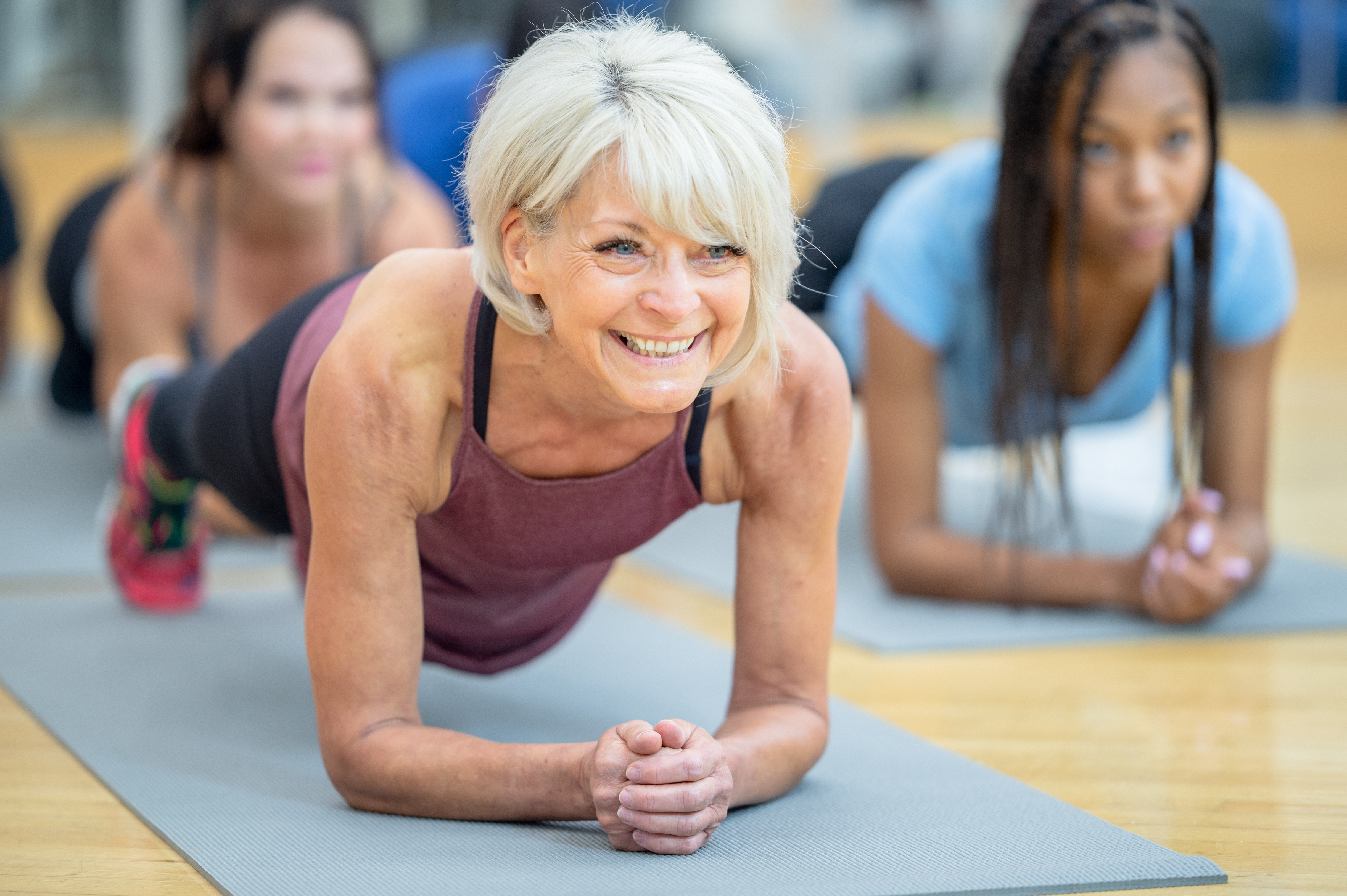 A fit and athletic senior Caucasian woman is participating in a multi ethnic group fitness class at a gym. She is holding a plank position on her yoga mat. She is smiling and happy.