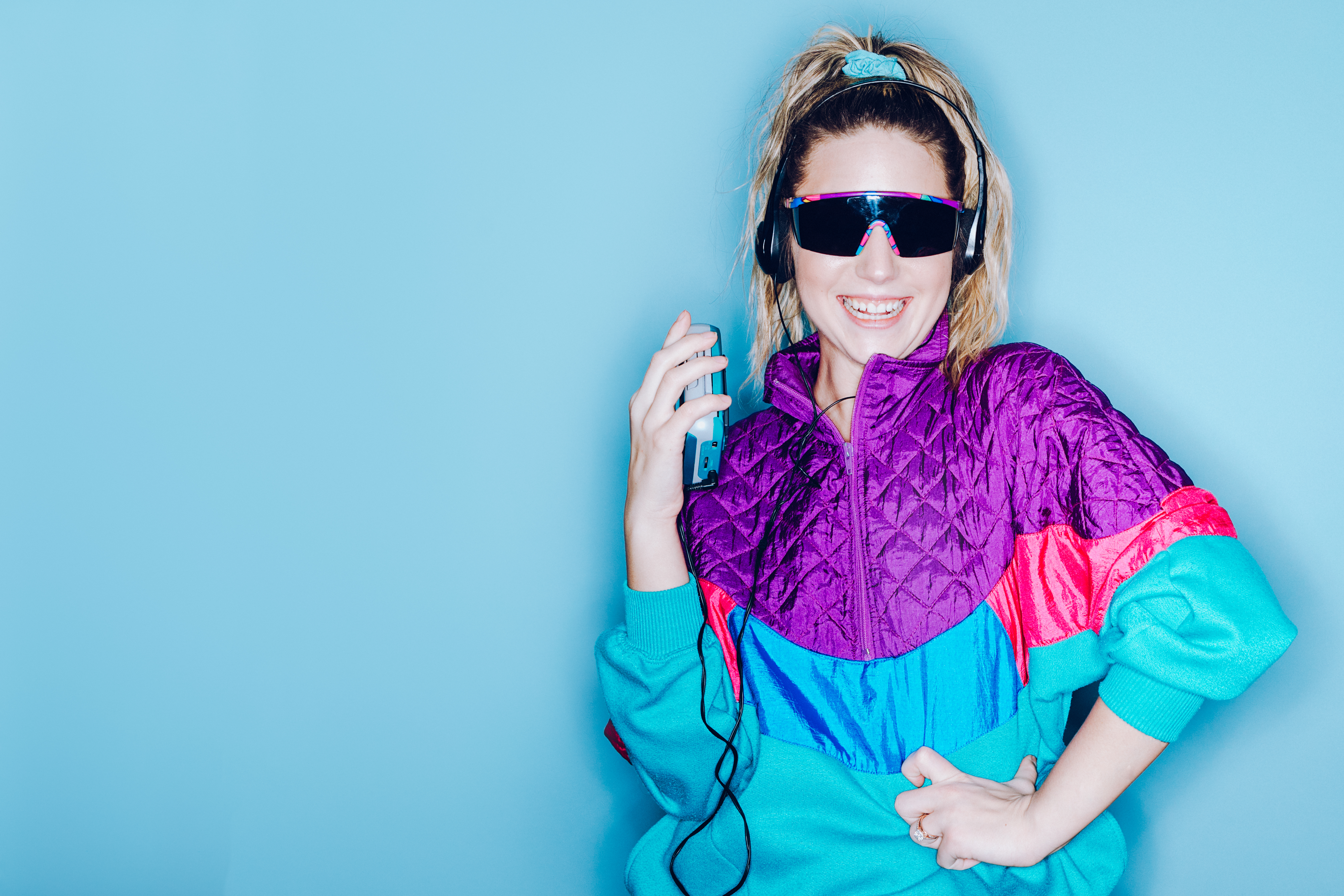 A woman wearing clothing styled after the 1980's and 1990's listens to music on her personal cassette tape player in front of a large bright blue background.  Shot with a ring flash.