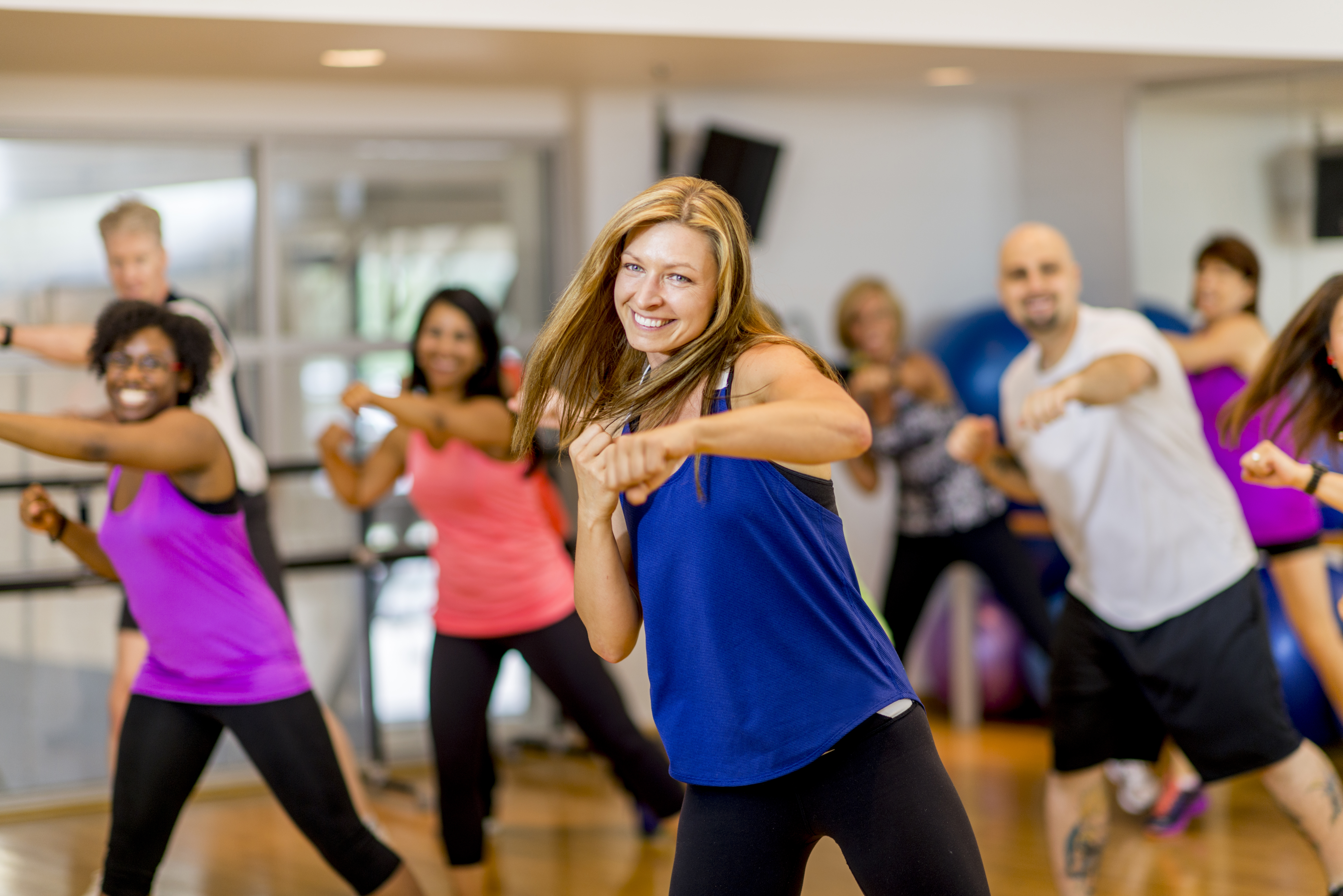 A woman is leading a multi-ethnic boxing class, everyone is wearing athletic clothing and is throwing a punch together - they are smiling and looking at the camera.