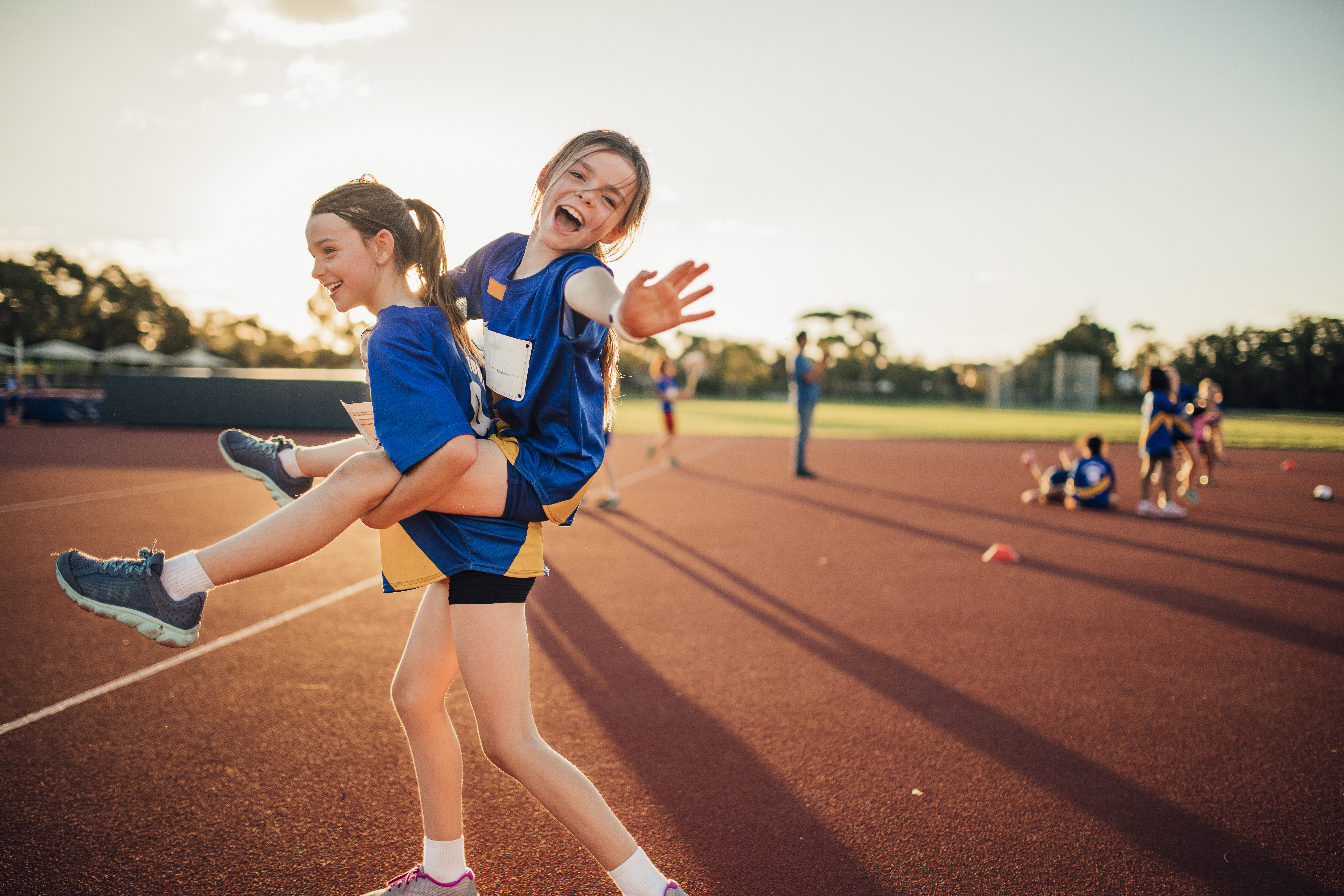 Two girls are messing around at athletics club on the running track. One is giving the other a piggy back and she is reaching out for the camera.